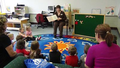 Children's services librarian reads to children during a storytime session. Children sit in a half circle on the floor.